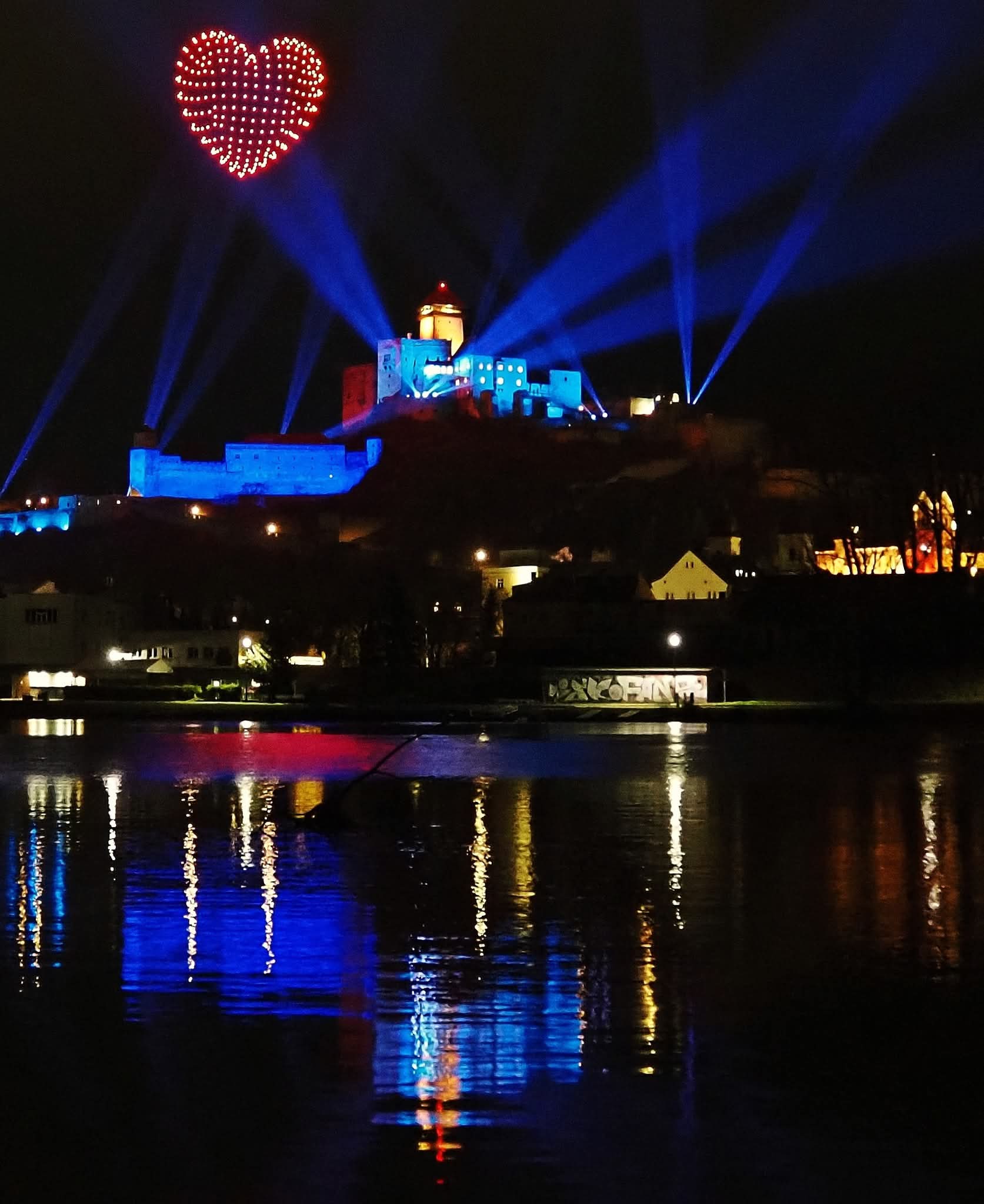 Red heart formed by drones in the night sky above Trenčín Castle with blue laser beams and reflections in the river below
