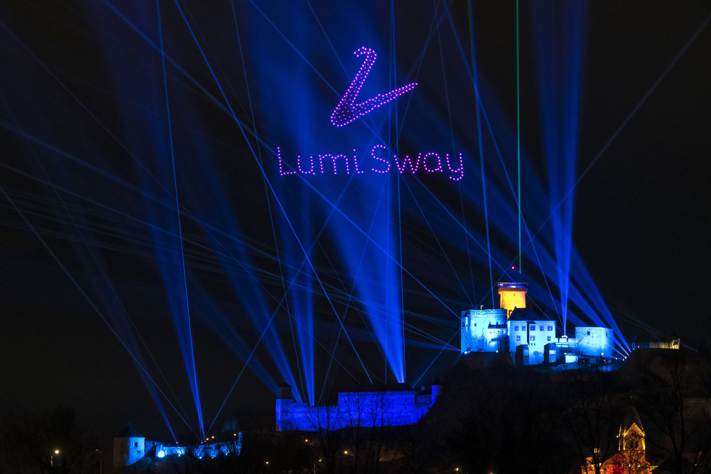 Lumi Sway logo and brand name formed by illuminated drones above Trenčín Castle with blue laser beams